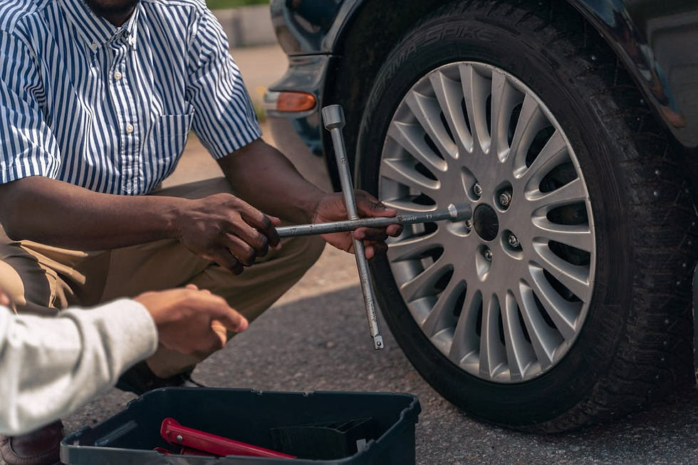 Two people change a car tire using a wrench. One wears a striped shirt, the other reaches from a toolbox. They're outdoors on pavement.