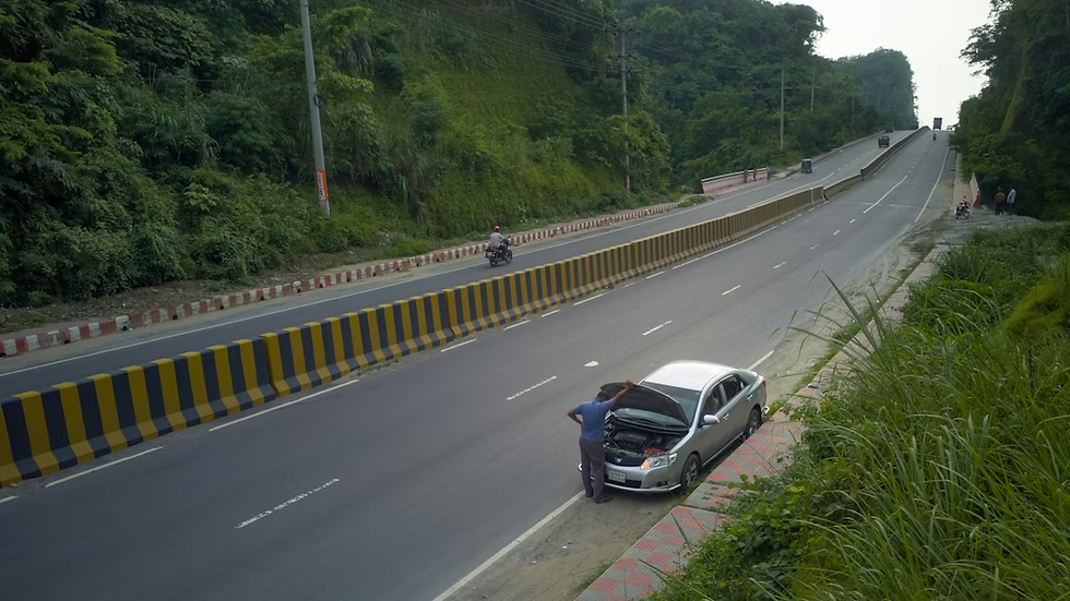 Man checks engine of a silver car on roadside, separated by a striped barrier. Motorbike rides by. Lush greenery surrounds, creating a calm setting.