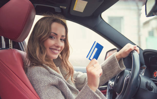 Smiling woman in a red car seat holds up a blue card while driving, with a bright car interior.