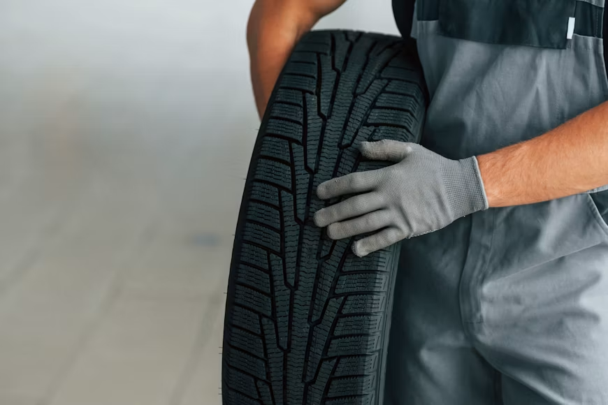 Gloved mechanic holds a black car tire in a workshop, checking its tread against a gray background.
