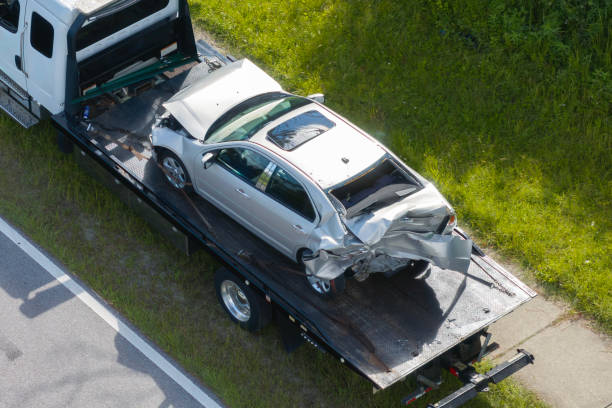 A damaged silver car on a tow truck's flatbed, set against a grassy roadside. The car's rear is heavily dented, indicating a crash.