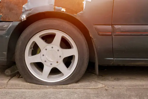 Flat tire on a dark car parked on pavement, with a damaged wheel arch and warm sunlight glare.