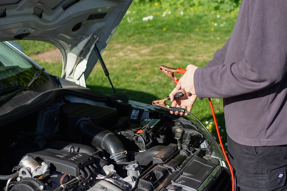 Person holding jumper cables near a car battery under an open hood on a sunny grassy roadside