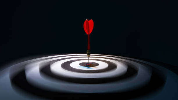 A red dart hits the bullseye on a black and white concentric target, surrounded by a dark background, symbolizing precision and focus.