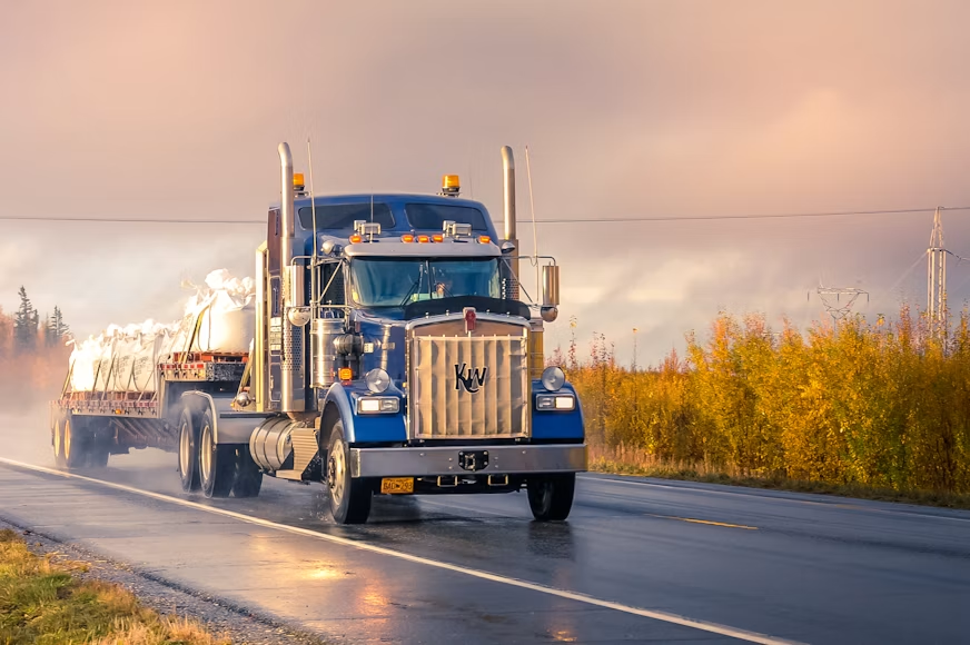 Blue Kenworth semi truck with KW grille hauling tarped cargo on a wet highway at sunrise, autumn trees glowing beside it.