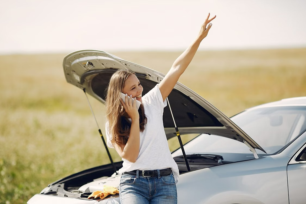 Smiling woman on a phone call beside a car with its hood open in a grassy field, raising her arm in relief or triumph.