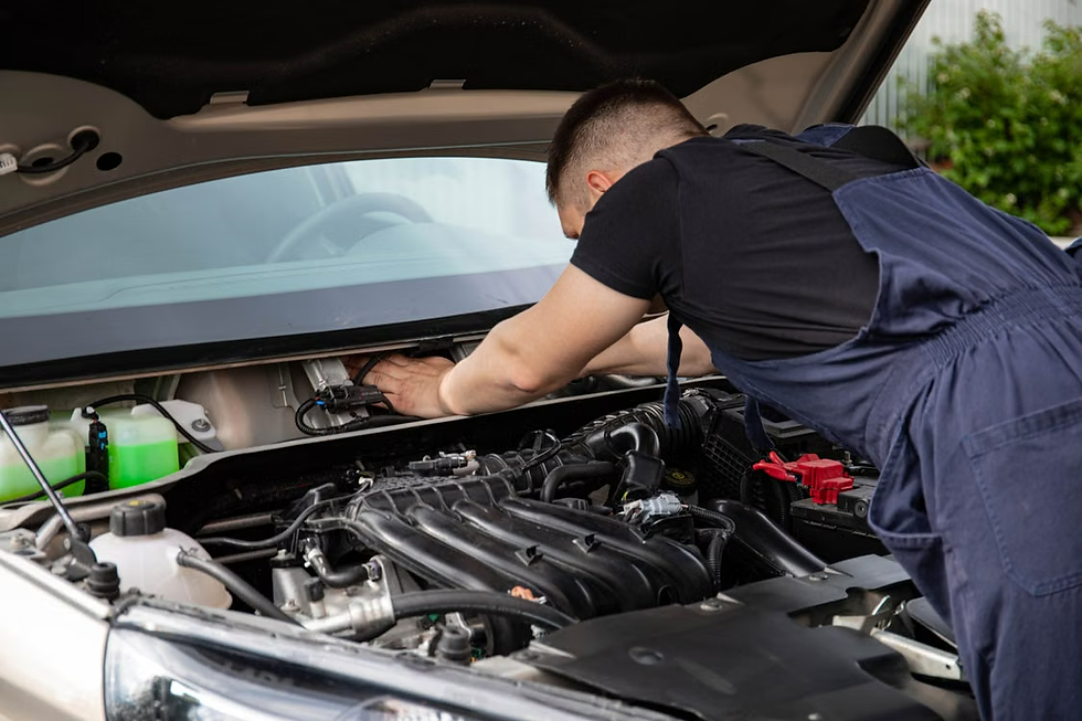 Mechanic in blue overalls works under an open car hood, leaning into the engine bay outdoors.