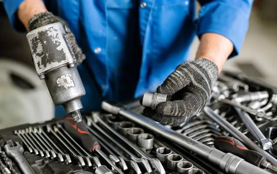 Mechanic wearing gloves and a blue uniform holding a power tool and socket above a toolbox filled with wrenches and tools in a workshop.