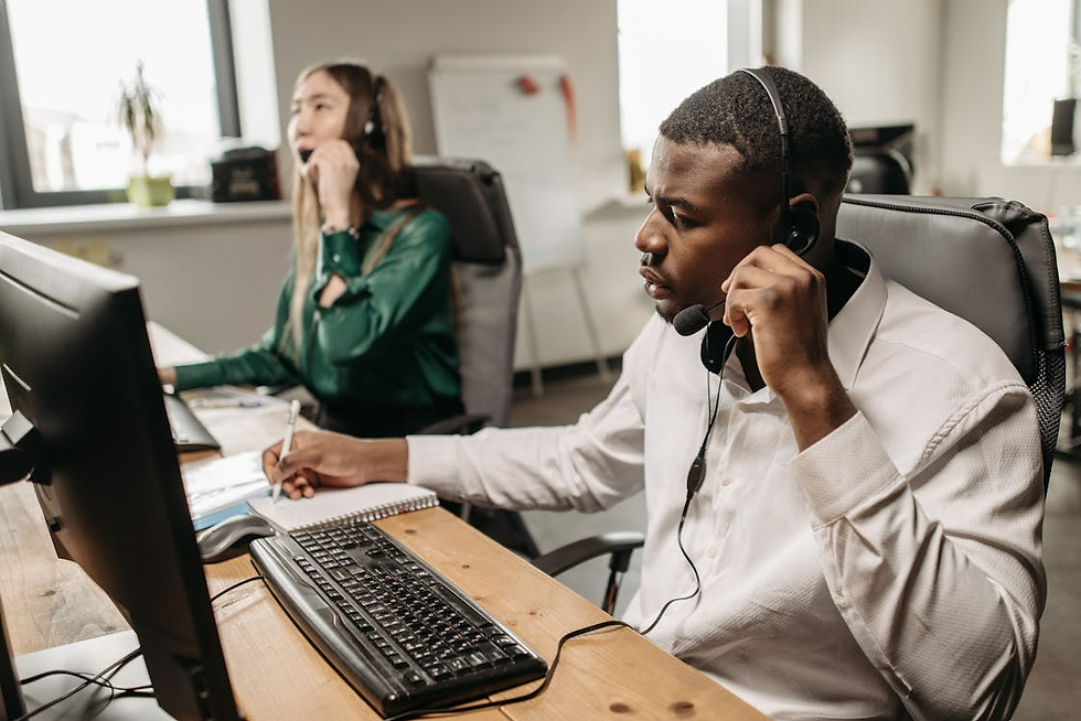 Two people in an office wearing headsets. One writes notes; the other talks. Computers and notepad are on a wooden desk. Daylight from windows.