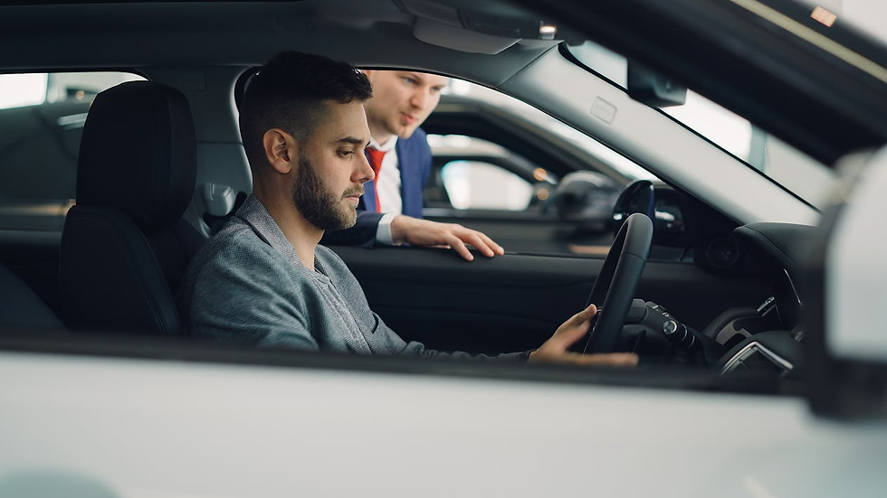 Man in gray blazer sits in car, hands on wheel, focused. Another man in suit leans in from side, discussing. Car showroom background.