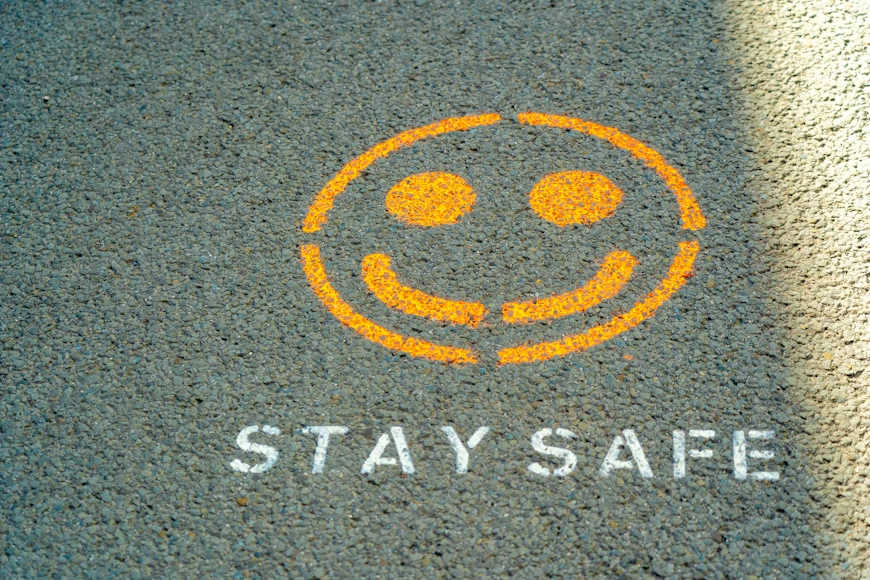 Smiley face and "Stay Safe" text stenciled in orange and white on asphalt. A shadow partially covers the design, creating a thoughtful mood.