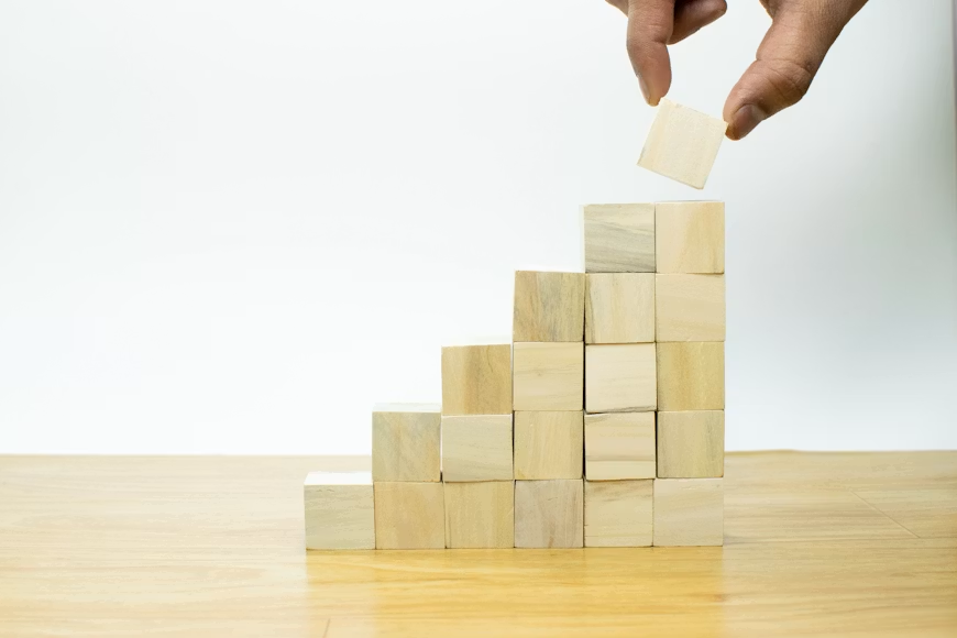 Hand placing a wooden block atop a staircase of blocks on a wood table, symbolizing growth against a white background.
