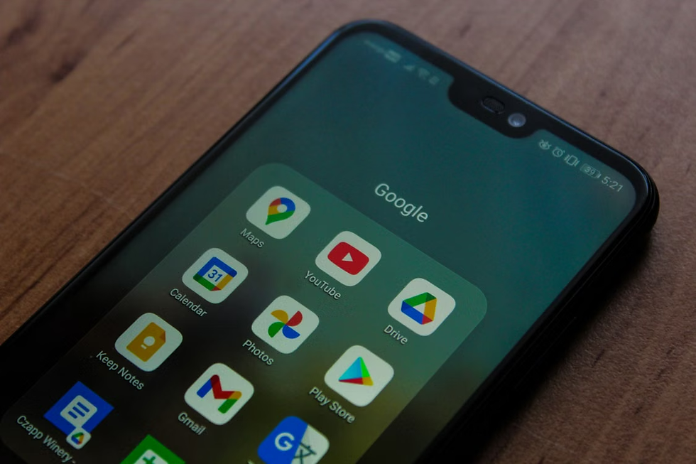 Smartphone on a wooden table showing a Google app folder with Maps, YouTube, Drive, Calendar, Photos, Gmail, and Play Store icons.