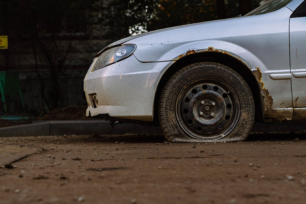 Close-up of a silver car with a flat, rusty front wheel on a muddy road. The car shows signs of rust and wear. Background is dimly lit.