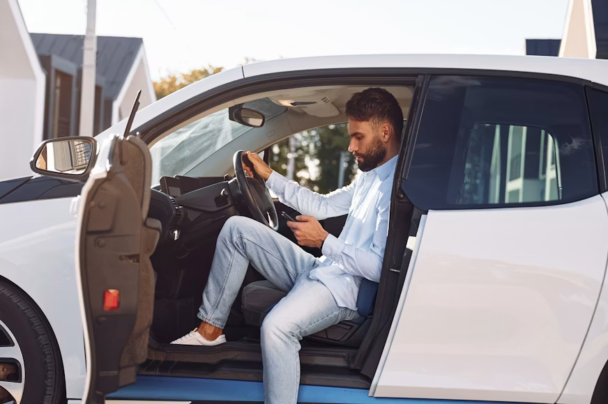 Man in a white car with the driver door open, checking his phone and holding the steering wheel in a sunny driveway.