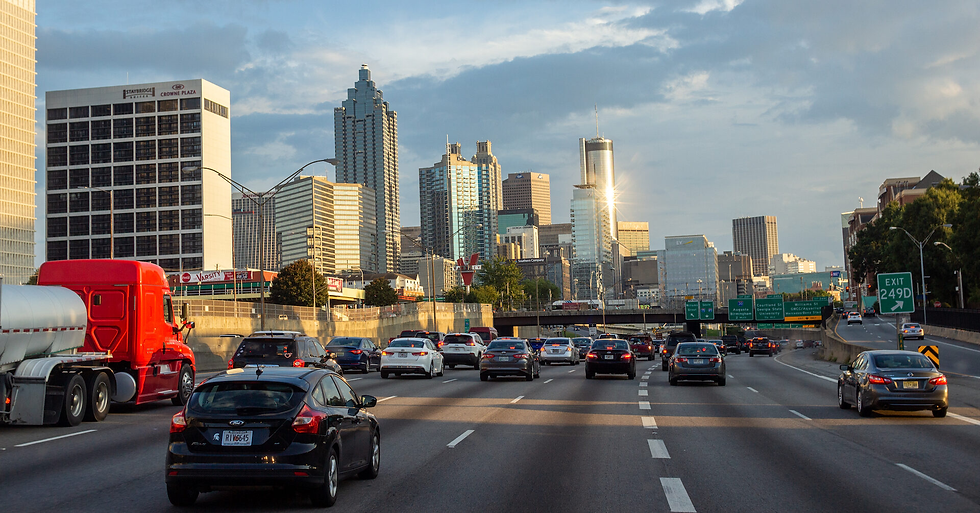 Cars drive on a highway towards a city skyline at sunset. A red truck is notable. Street signs and skyscrapers are visible.