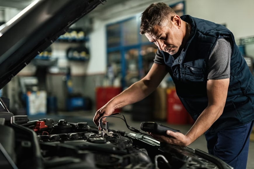 Mechanic in a blue vest uses diagnostic tool on car engine in a garage. Background shows blurred tools and shelves. Focused expression.