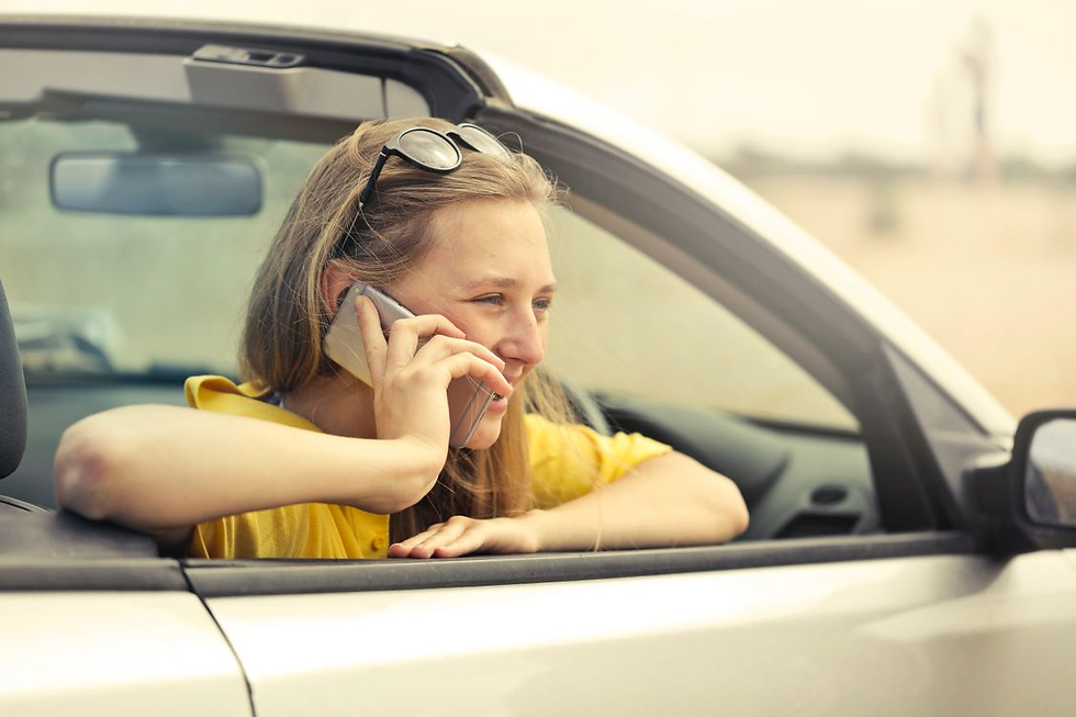 Smiling woman in a yellow shirt leans from a convertible window, talking on a smartphone in bright sunshine.