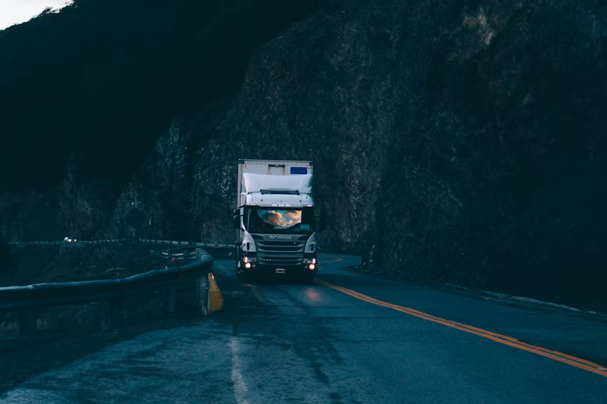 White Scania truck drives on a wet winding mountain road through a dark canyon, headlights glowing.