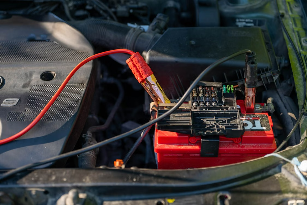Car engine bay with a red battery connected by jumper cables and a fuse box, showing a repair or jump-start setup.