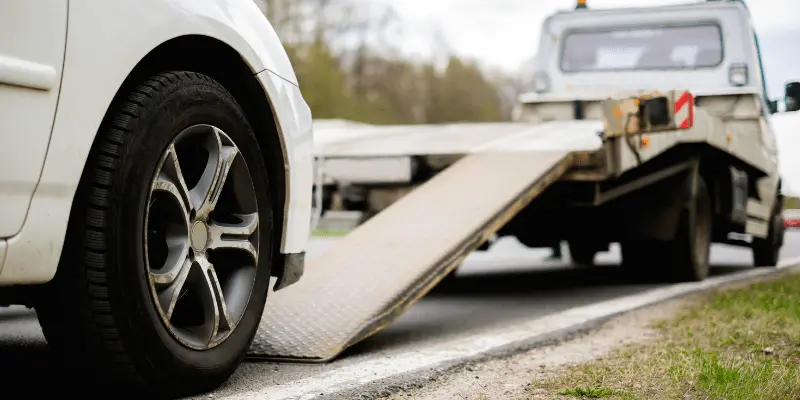 Car at roadside with tire visible, ready to be loaded onto a tow truck's ramp. Overcast day with trees in the background.