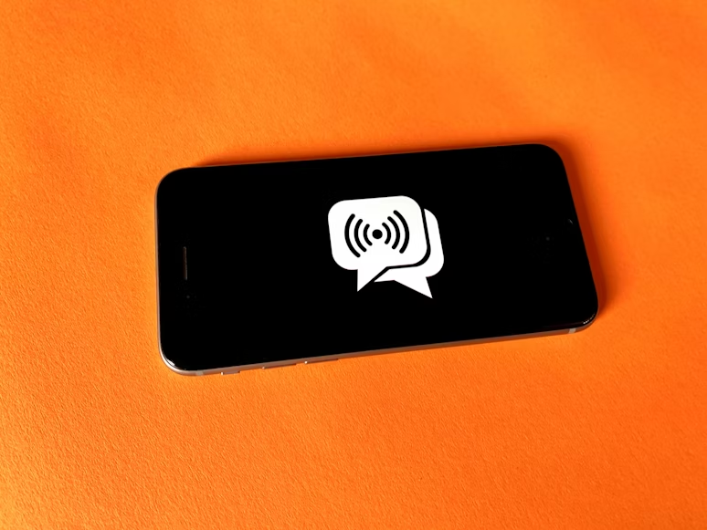 Smartphone on orange surface displaying white chat bubble with broadcast symbol, evoking communication and connection themes.