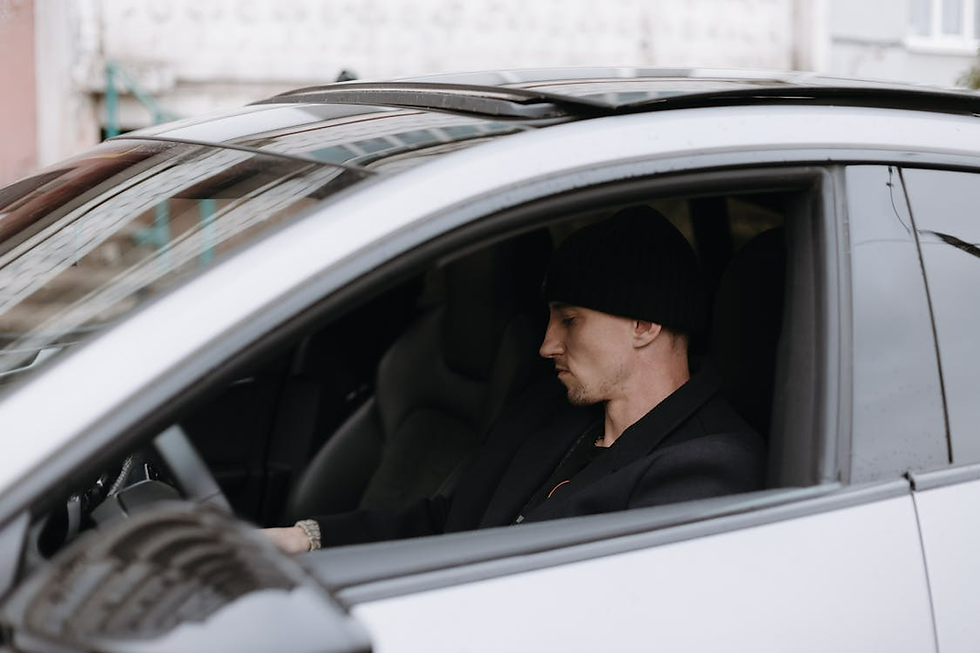 Man in black beanie sits in silver car, looking down. Urban background outside window. Calm mood.