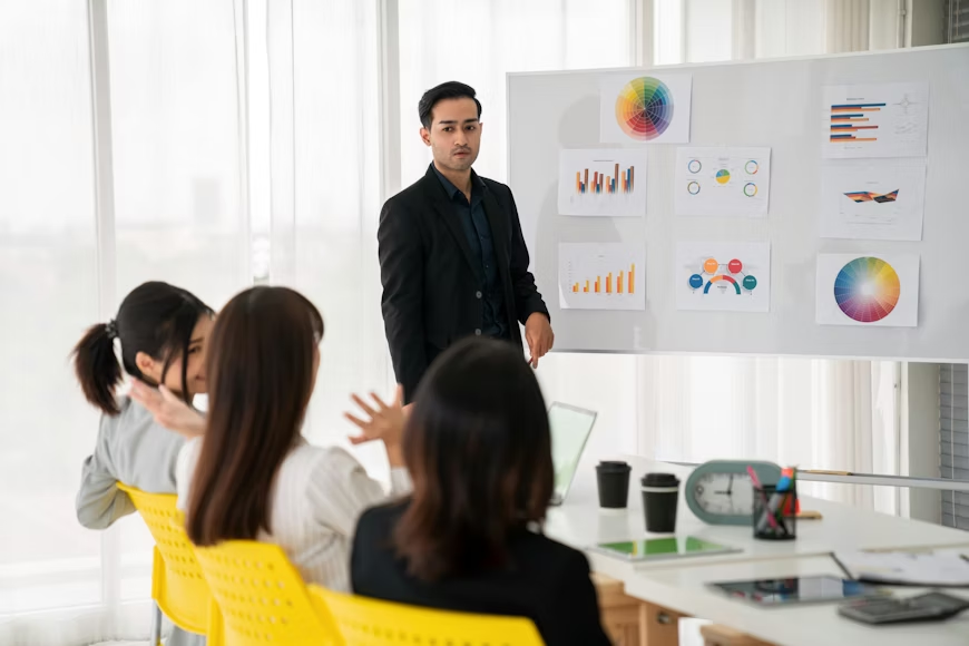 A man in a black suit presents graphs on a whiteboard to seated colleagues in a bright office. Yellow chairs, laptops, and charts visible.