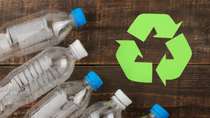A wooden table with five empty plastic bottles with either a white or blue lid. There are three green arrows in the shape of the recycling symbol.