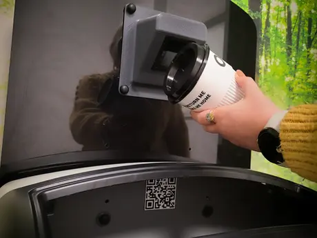 A person wearing a yellow jumper is scanning a coffee cup at a reusable drop off point. There is forest wall paper in the background.
