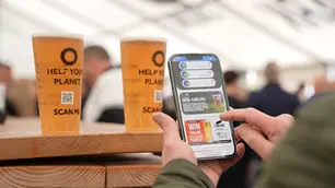 Two reusable cups filled with beer, and a man holding a phone displaying Circulayo's AppNostic with environmental impact.