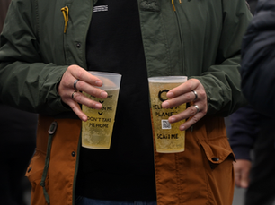 A man wearing a duotone coat in green and orange is holding two reusable cups filled with beer in his hands.