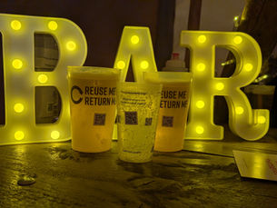 A bar sign lit up with bulbs with three reusable cups filled with beer and lemonade. These cups have messaging regarding reuse and returns, and QR codes are also attached.