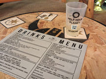 A cork table with Freedom Brewery drink mats, and a reusable cup that says "Help Your Planet" and a QR Code with "Scan Me". On the table there is a drinks menu.