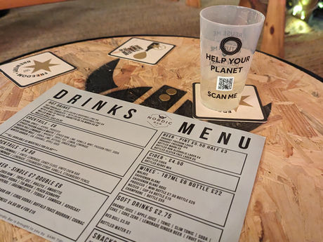 A cork table with Freedom Brewery drink mats, and a reusable cup that says "Help Your Planet" and a QR Code with "Scan Me". On the table there is a drinks menu.