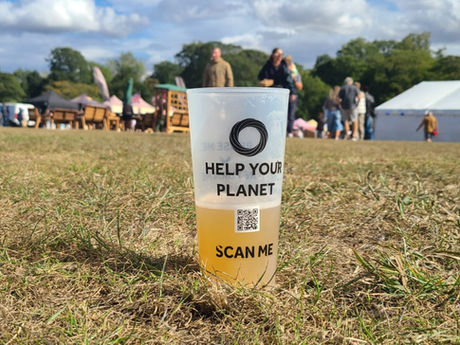 A reusable cup that is half-filled with beer. On the cup is a QR code and text that states "Help Your Planet" and "Scan Me". The reusable cup is placed on a festival field floor, with people and tents behind them.