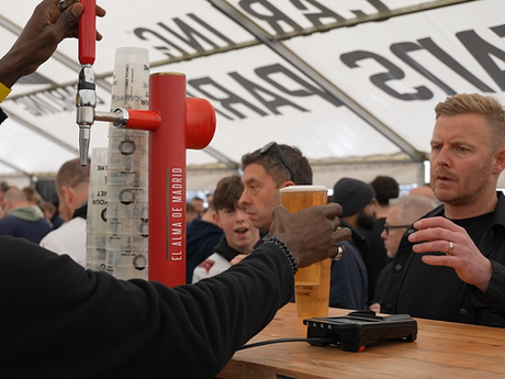 A member of bar staff handing over a reusable cup of beer to a customer. There is a stack of reusable cups by the pump. There is a crowd of people behind the bar.