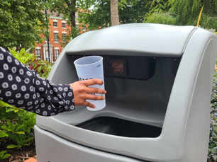 Someone is scanning a reusable cup at a bin and dropping it into the bin. The bin is grey, and behind it is foliage, such as trees and bushes. There is also a bush in the background.