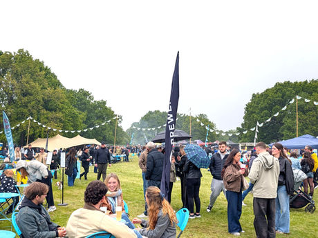 Lots of people gathered in a field with gazebos and umbrellas. People have reusable cups in hands.