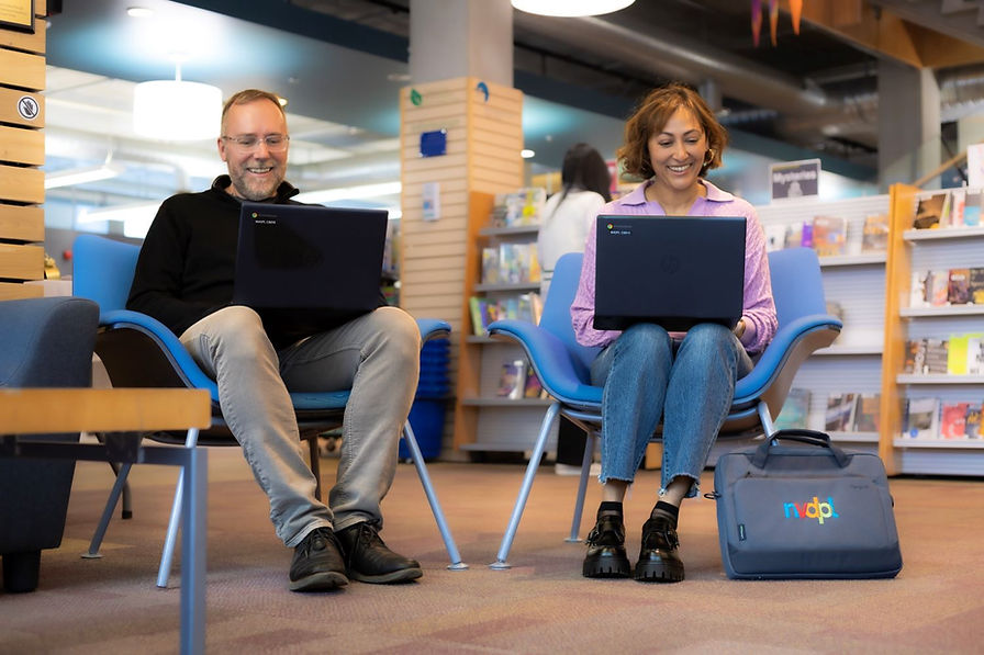 Two people sitting on chairs smiling while they look at their laptops.