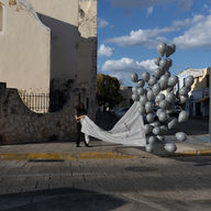Una mujer, en la calle, frente a una iglesia en Mérida, Yuc., sostiene una manta que se va elevando con muchos globos grises, durante el atardecer soleado.