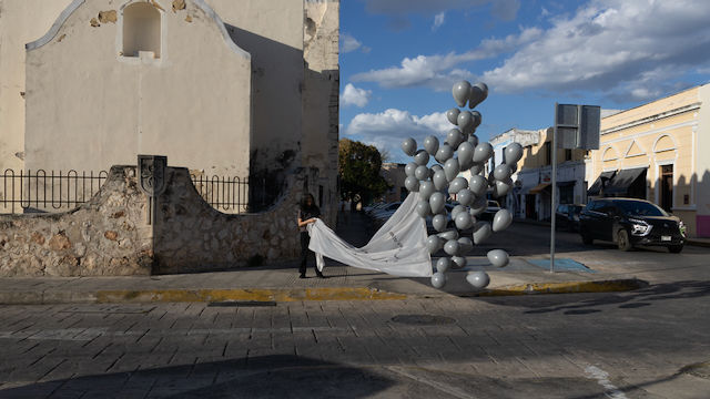 Una mujer, en la calle, frente a una iglesia en Mérida, Yuc., sostiene una manta que se va elevando con muchos globos grises, durante el atardecer soleado.