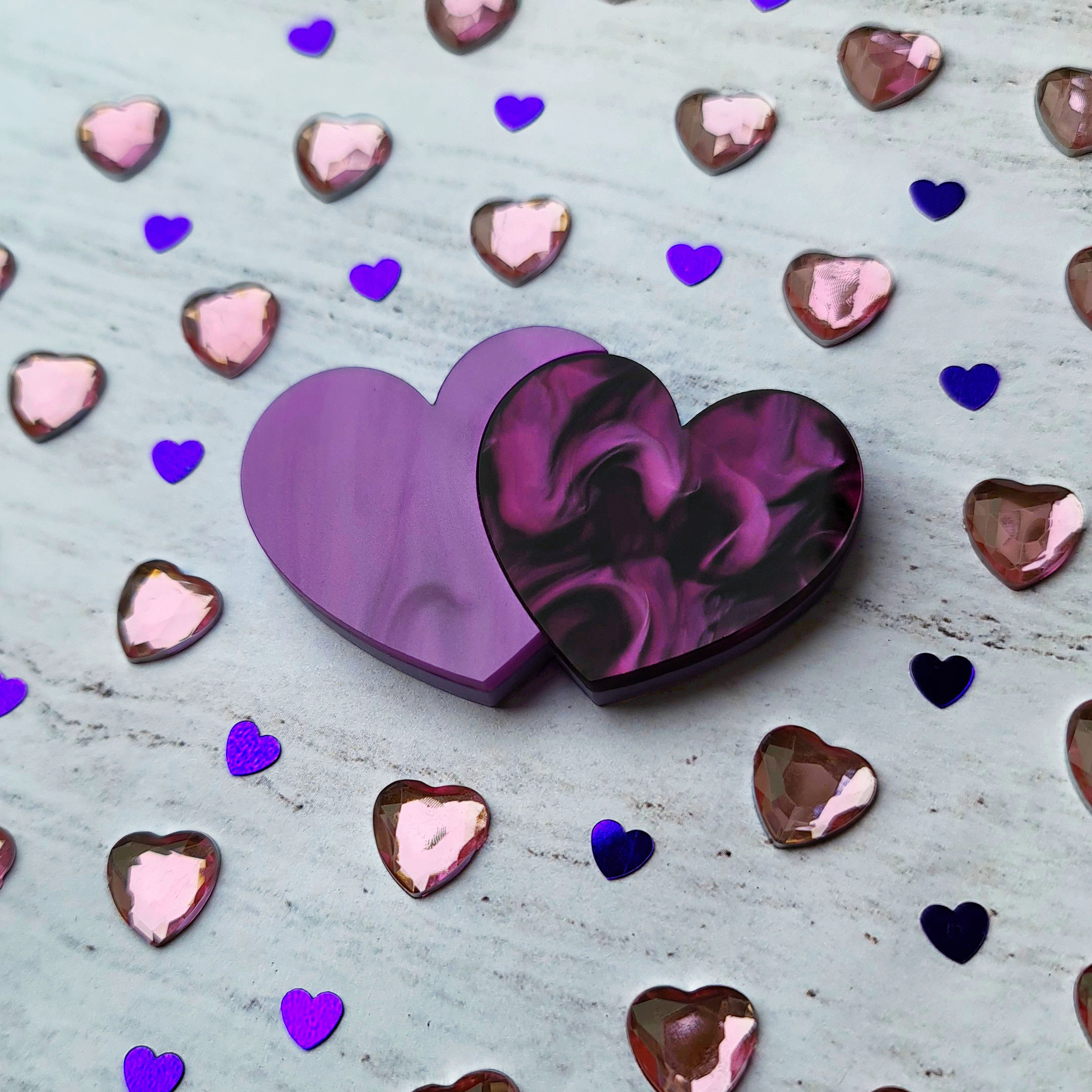 The Double Hearts brooch on a marble background surrounded by lilac heart gems