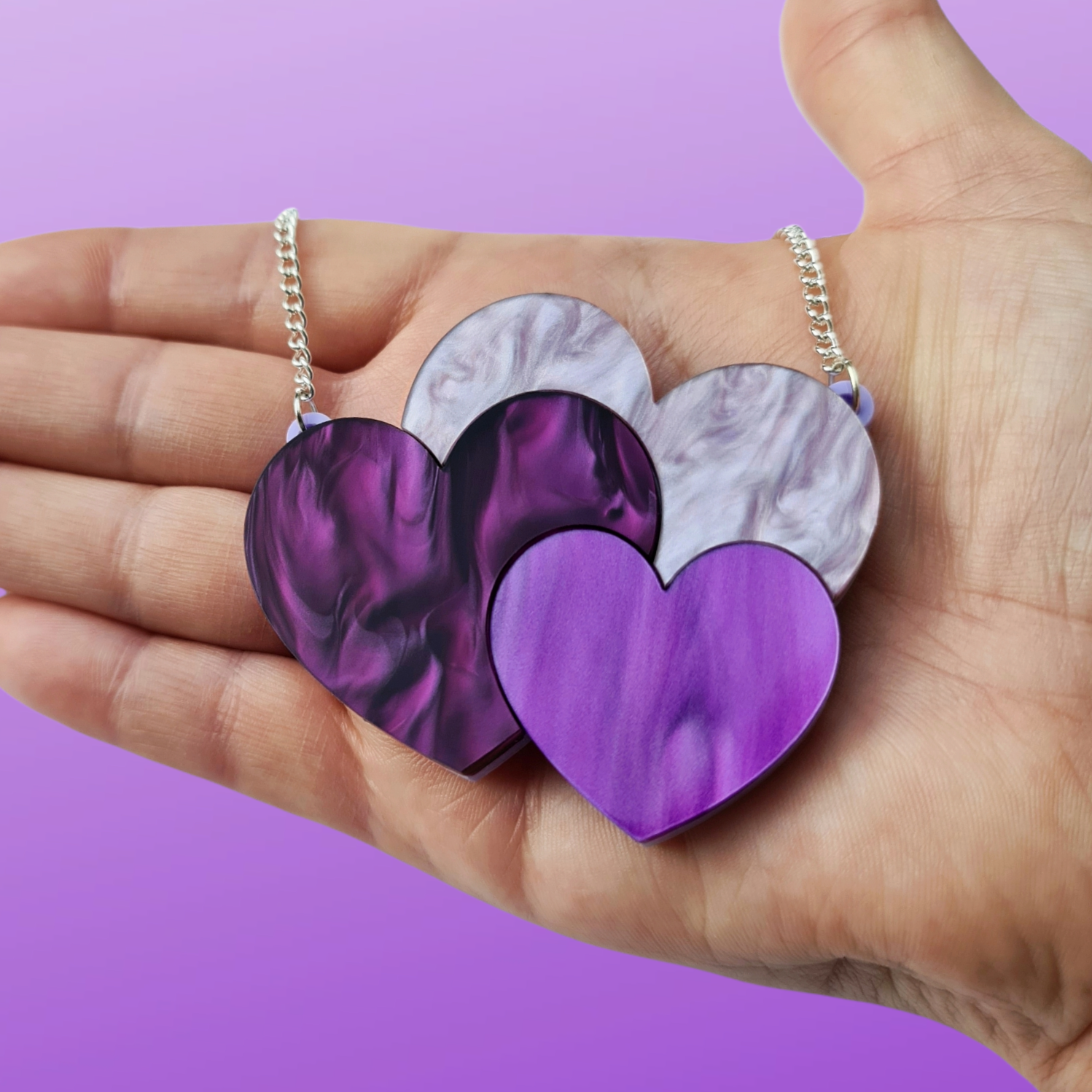 The Hearts Trio necklace in the makers hand in front of a lilac background, to show it's scale