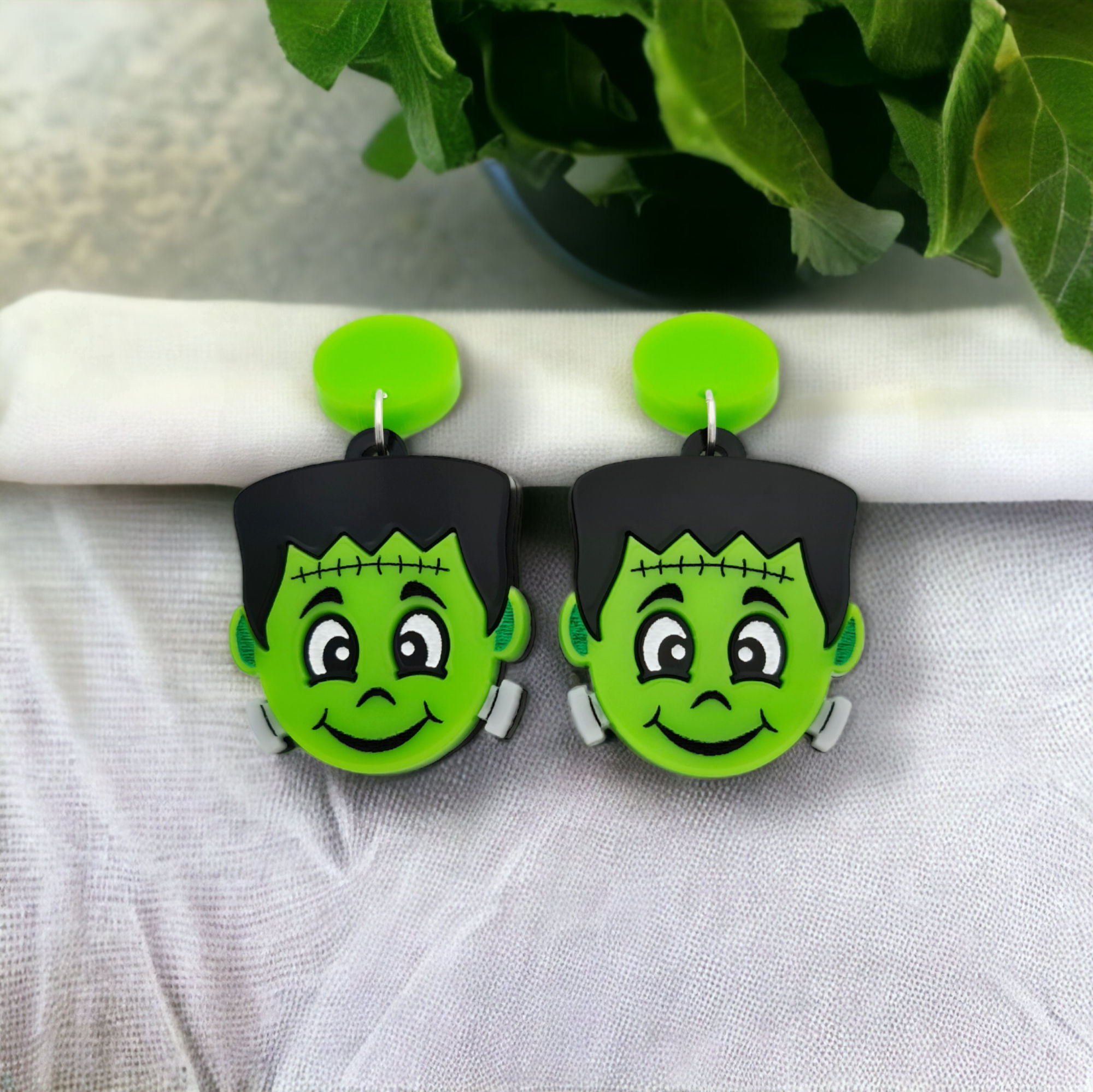 A friendly looking pair of acrylic Frankenstein earrings displayed on a white cloth background