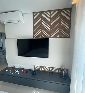 Na imagem temos um painel de TV de um quarto composto por um painel em madeira desenhado com frisos na diagonal e um painel em laca branca na horizontal. Na parte de baixo temos um aparador cinza escuro com gavetas e bibelôs em cima.