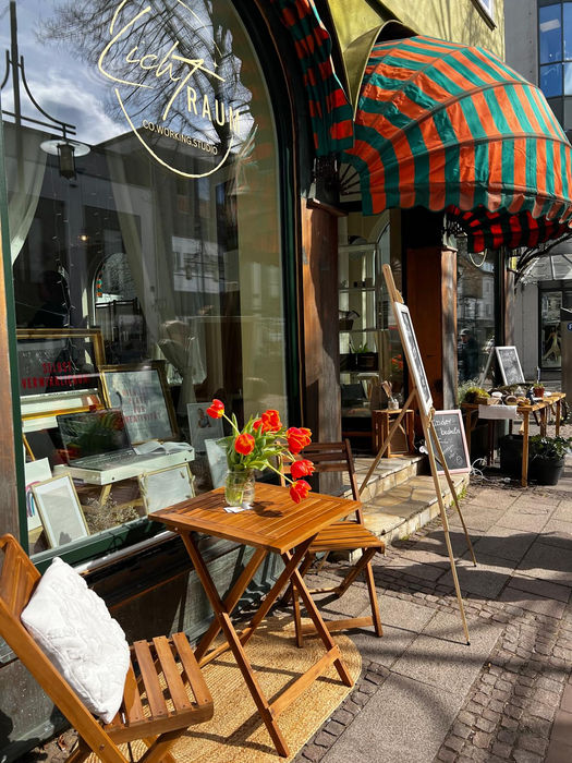 Außenansicht des Lichtraums mit großen Schaufenstern, bunten Markisen und einladend platzierten Stühlen vor dem Eingang.