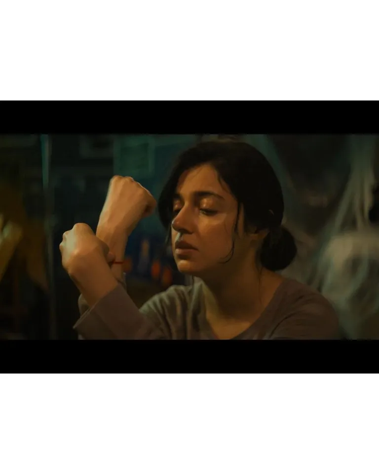Film still of a woman holding one wrist near her face in a dim interior.