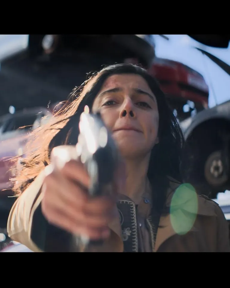 Film still of a woman aiming a handgun toward the camera, with a vehicle behind her.