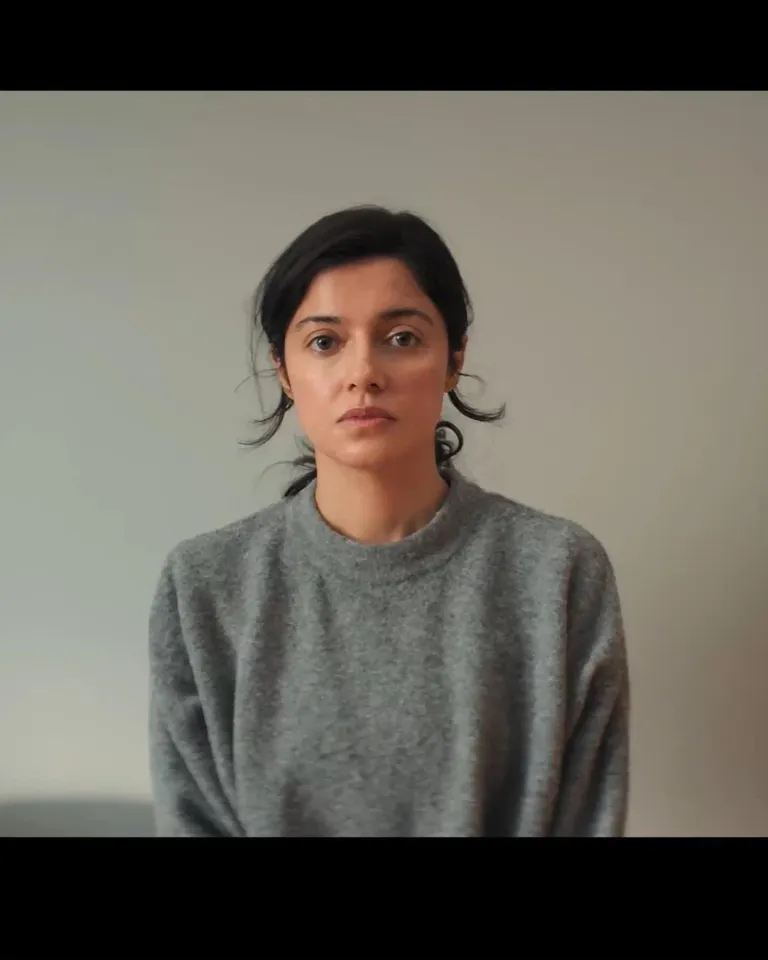Film still of a woman in a gray sweatshirt against a plain wall.